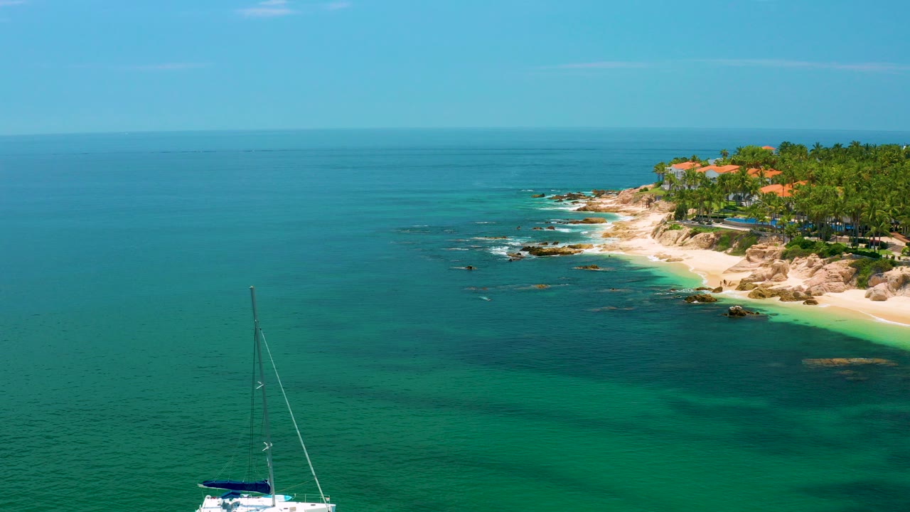 At Palmilla Beach, Baja California Sur, a sailboat sits near the lower left below blue-green water, rocky sand, palms, orange roofs, and no visible people.