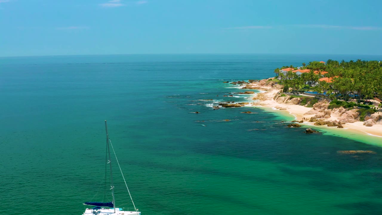 At 0:01, the sailboat shifts closer to the bottom edge while Palmilla Beach rocks, sand, palms, orange roofs, and no visible people remain in the background.