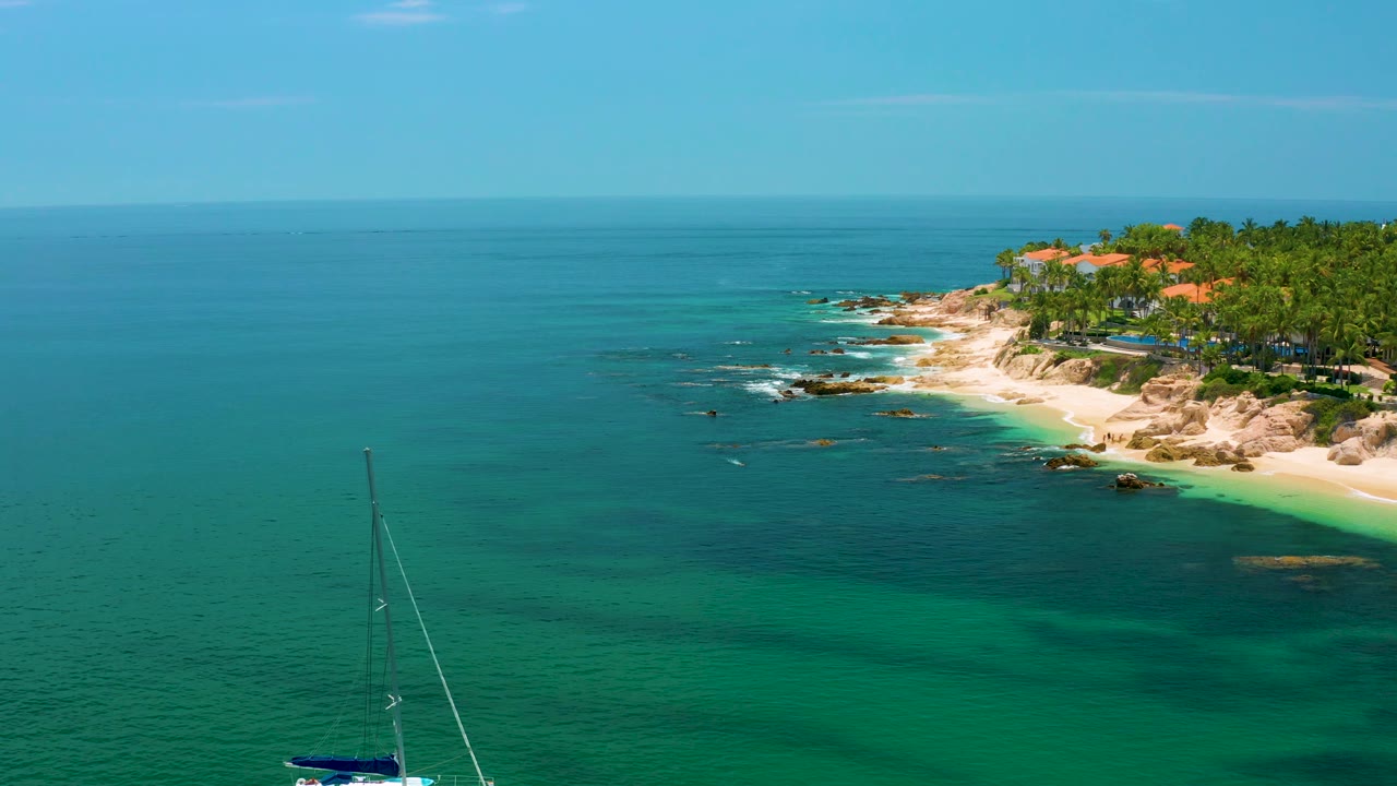 At Palmilla Beach, the sailboat sits partly cropped near the bottom while darker water patches, rocky shallows, palm trees, orange roofs, and no visible people are visible.