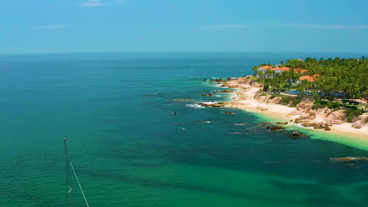 Palmilla Beach coastline from above, showing broad blue-green water in the foreground, a curved sandy beach, rocky outcrops, palm trees, distant roofs, and no visible people.
