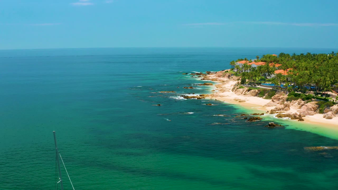 Aerial coastal frame at Palmilla Beach with clear blue water, a narrow bright sand strip, rocks breaking the surf, dense palms, red-roofed beachfront structures, and no visible people.