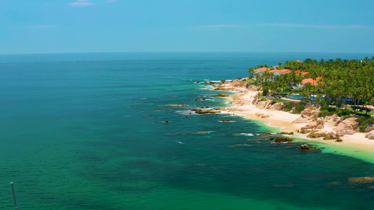 Wide aerial coastline with turquoise shallows, rocks, palms, and red roofs.