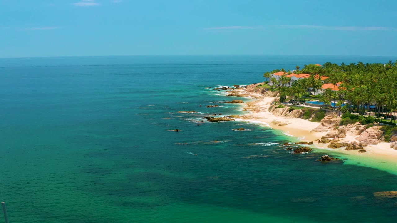Aerial frame of Palmilla Beach coastline with calm blue water across the foreground, a rocky point, pale sand, palm trees, coastal buildings, and no visible people.