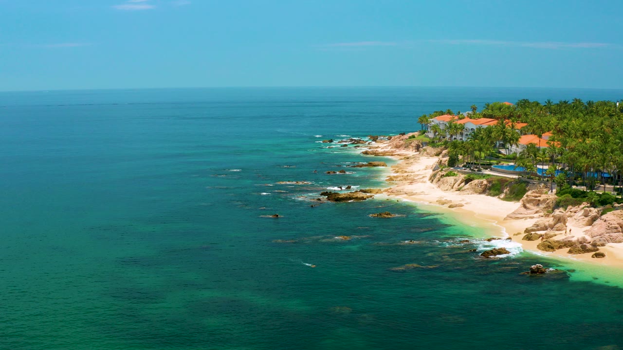 Aerial view over Palmilla Beach in Baja California Sur, with turquoise water, pale sand, shoreline rocks, palms, red-roofed buildings, and no visible people.
