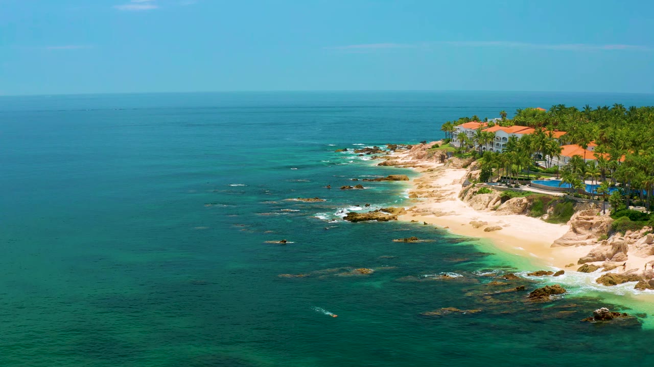 Wide aerial coastline view with rocky shallows, pale sand, palms, and resort-style buildings.
