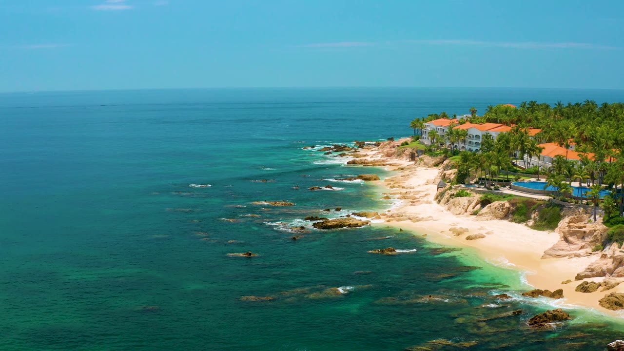 Aerial view of Palmilla Beach with the resort complex near the upper right, a curving sandy shoreline below it, scattered rocks offshore, and tiny people on the beach.