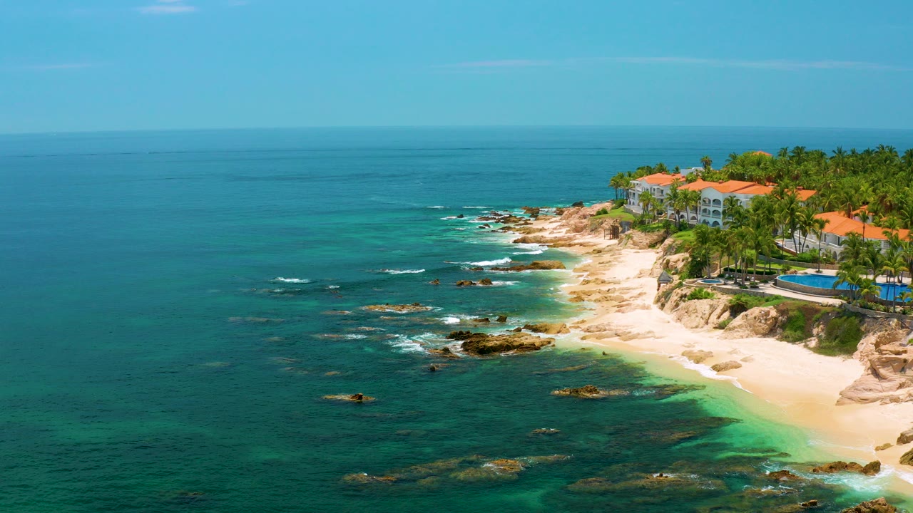 Palmilla Beach in Baja California Sur seen from above, with deep blue water at left, pale sand and rocks along the shore, palm trees and red-roof buildings at right, and no visible people.