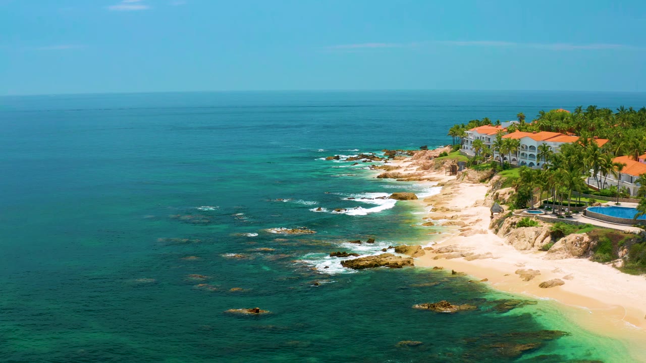 Aerial frame over Palmilla Beach showing more open water at left, scattered rocks in the surf, the sandy cove, resort roofs, palm trees, and no visible people.
