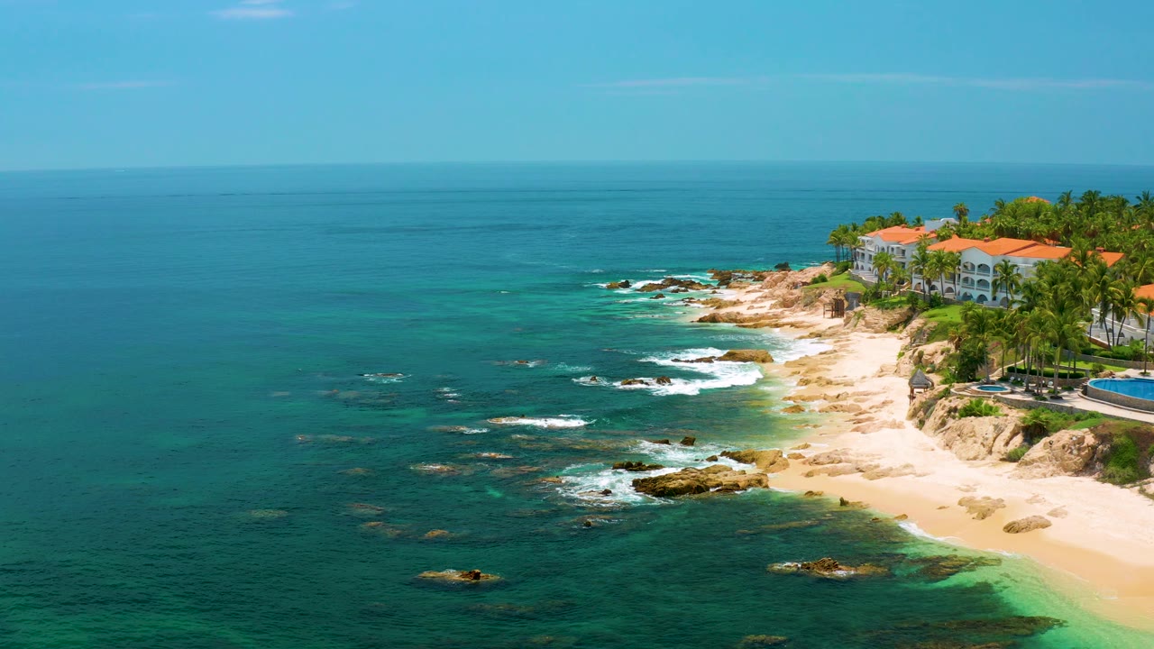 Mid-clip view of Palmilla Beach with broad teal ocean foreground, small offshore rocks, surf tracing the sand, cliffside resort buildings at right, and no visible people.