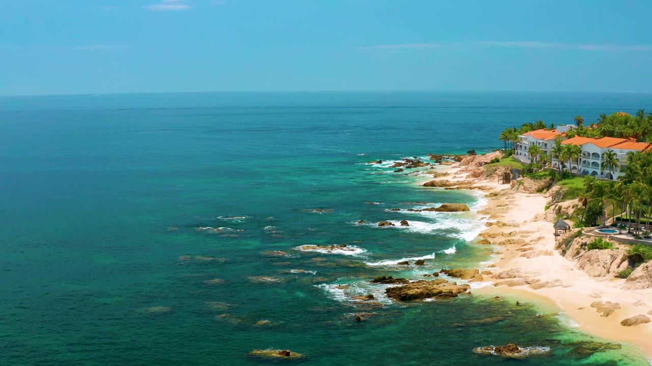 Palmilla Beach aerial frame showing deep blue ocean across the left, scattered rocks with small white wave breaks, a sandy cove, red-roofed buildings at upper right, and no visible people.