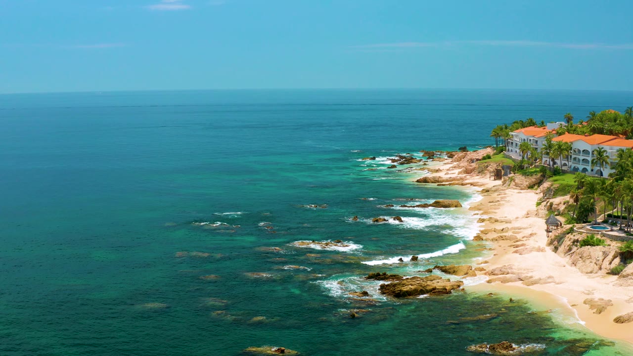 Aerial view of Palmilla Beach with more open water across the frame, white surf tracing the rocks near shore, red-roofed buildings partly cropped at the right edge, and no visible people.