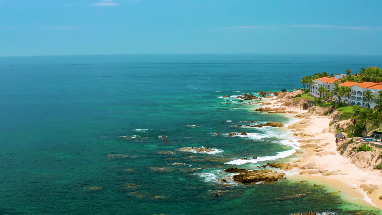 High coastal view with blue sea, beach, rock patches, surf, palms, and red roofs.