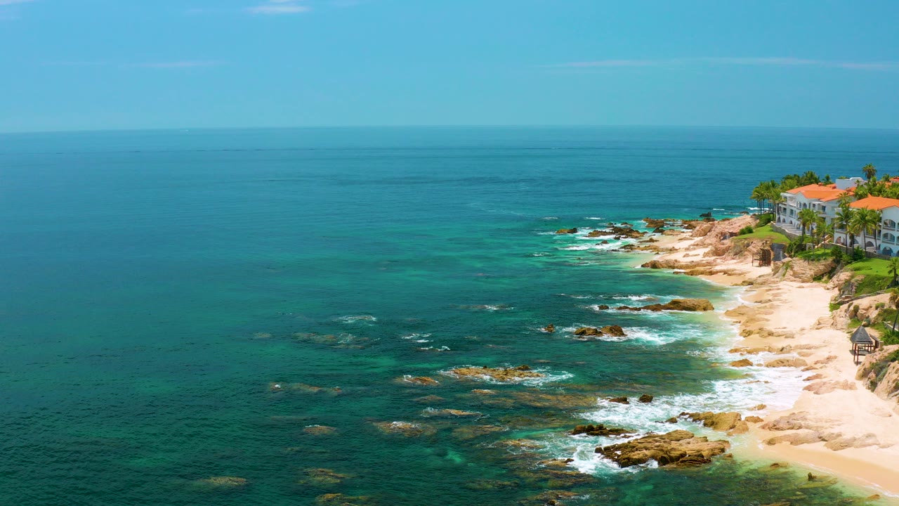 Opening aerial frame over Palmilla Beach, Baja California Sur, Mexico, with turquoise water, scattered rocks, pale sand, orange-roofed coastal buildings at the right edge, and no visible people.