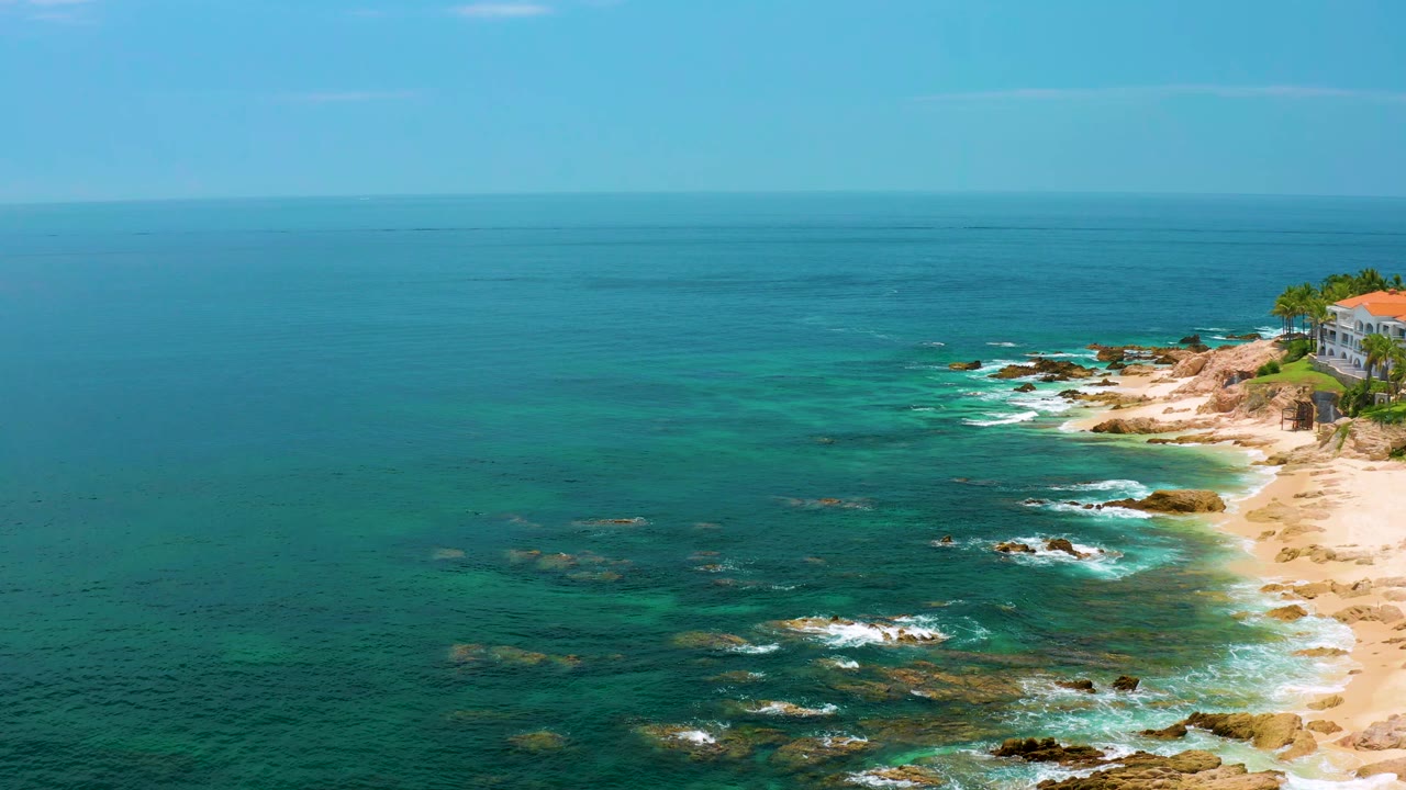 Aerial view at Palmilla Beach with the horizon high in frame, open blue sea across the left, white waves around nearshore rocks, and no visible people.