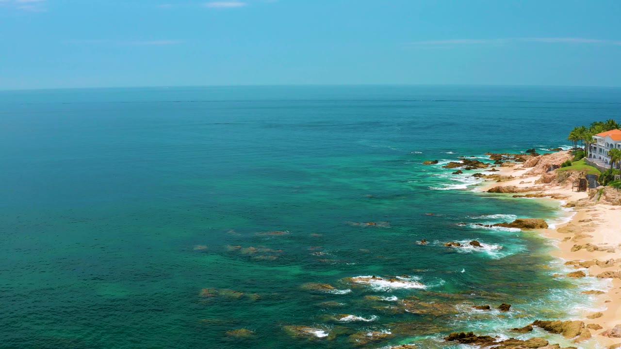 Palmilla Beach aerial frame showing turquoise water over submerged rocks, white surf near the sandy edge, distant blue sea, and no visible people.