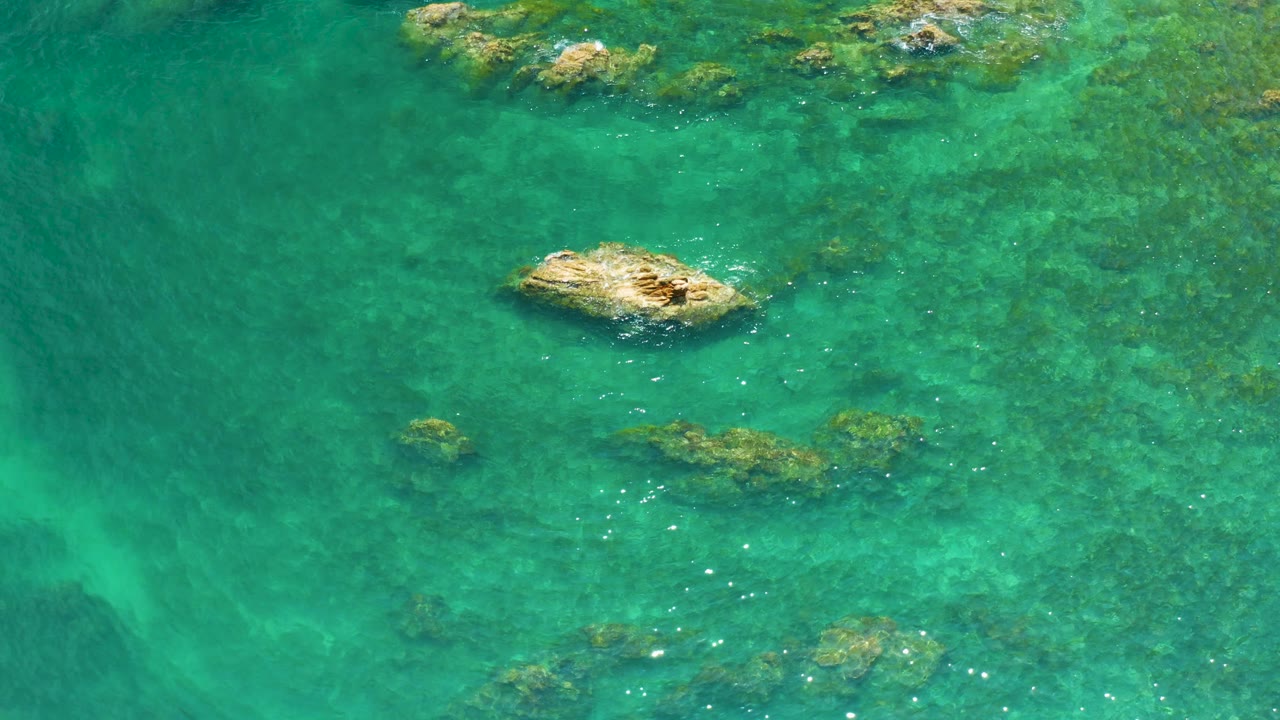 Overhead frame at Palmilla Beach showing a tan rock left of center, reef bands under green water, and no visible people or boats.