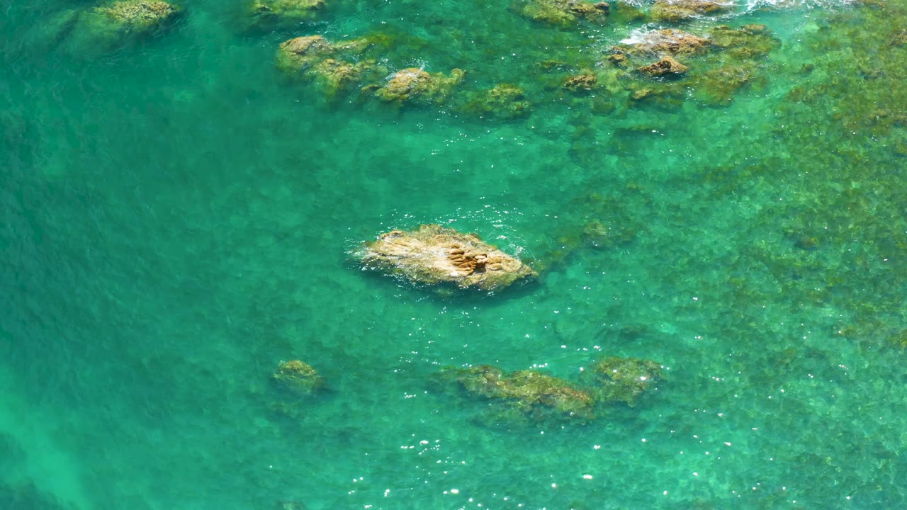 Aerial frame from Palmilla Beach showing the central tan rock shifted slightly lower in the composition, blue-green water over darker submerged patches, and no visible people.