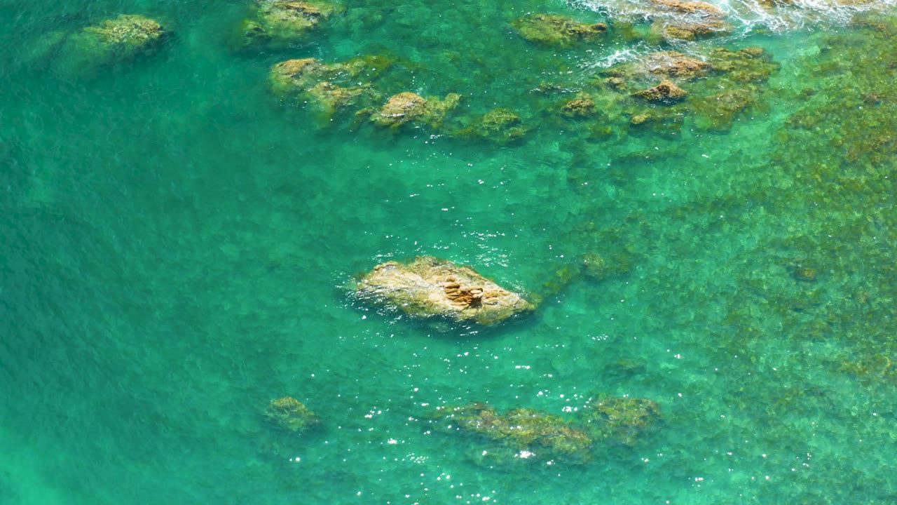 Palmilla Beach aerial still with the larger tan rock lower in frame, scattered submerged rocks across the right side, darker open water on the left, and no visible people.