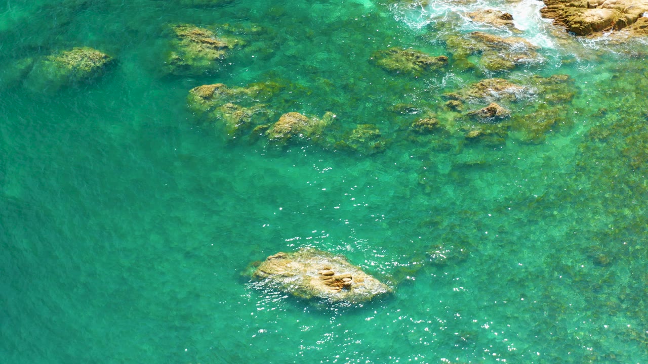 Overhead view of turquoise water at Palmilla Beach, Baja California Sur, with submerged tan rocks, surface sparkle, and no visible people.