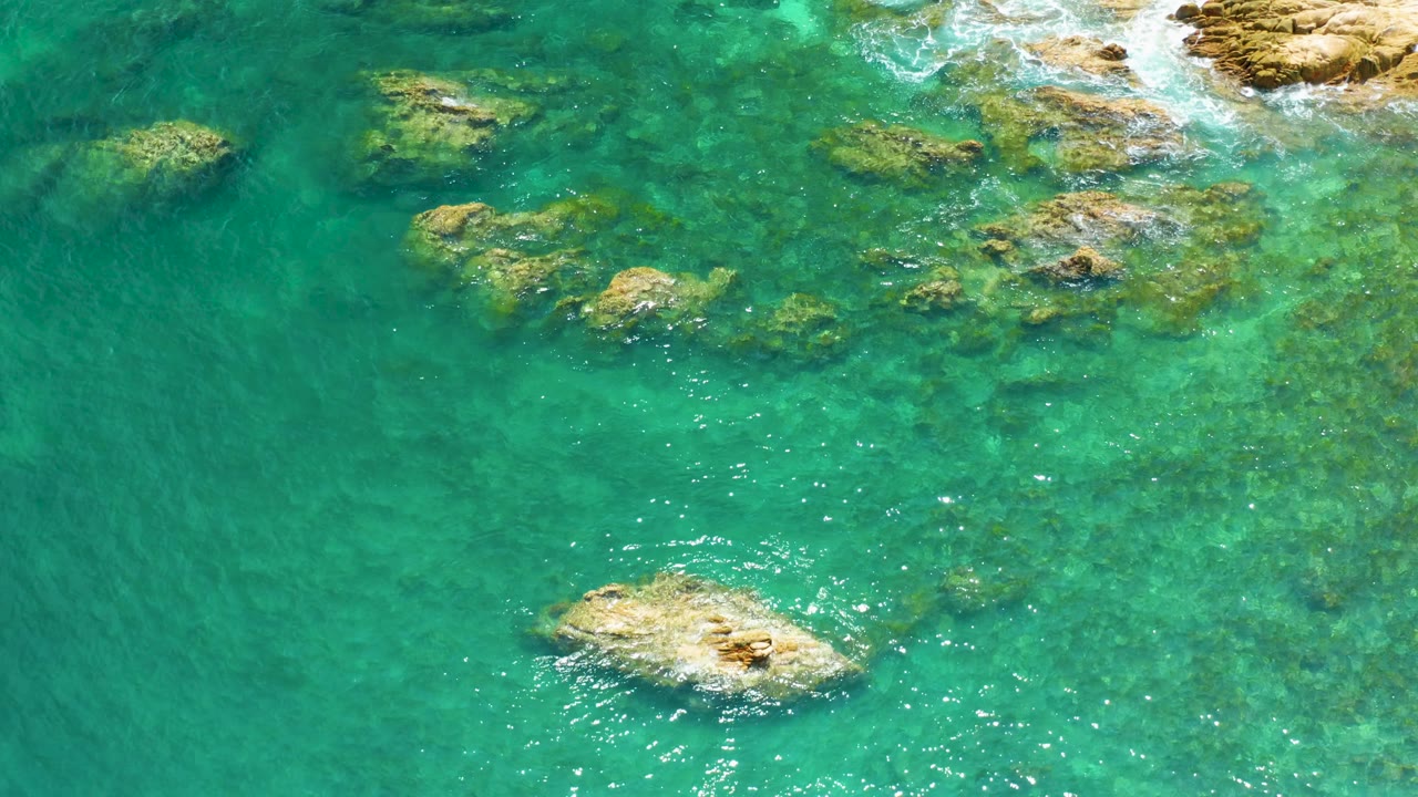 Overhead Palmilla Beach frame with transparent turquoise water, scattered underwater rocks, a bright tan rock low in frame, upper-right foam, and no visible people.