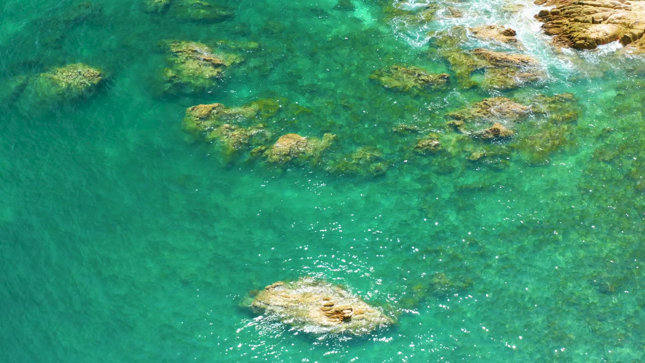 Mid-clip overhead view at Palmilla Beach showing open turquoise water on the left, submerged rocks toward the top and right, surface shimmer, and no visible people.