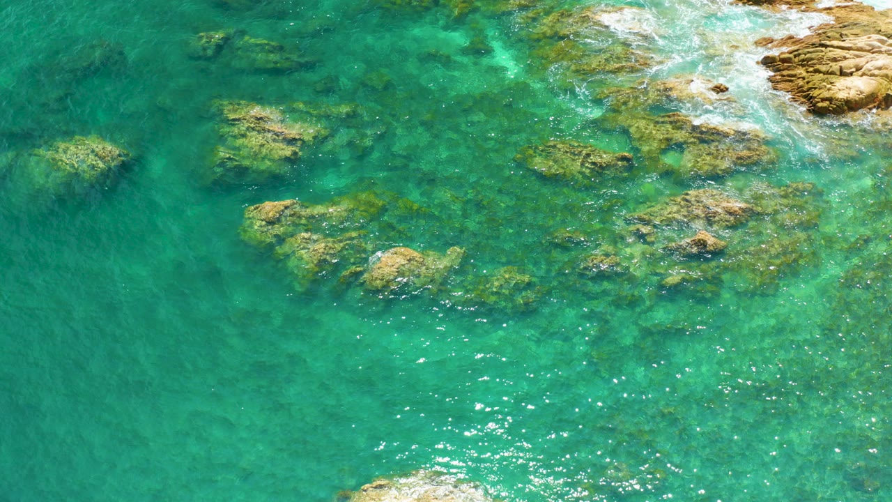 Overhead view of turquoise water and scattered reef rocks at Palmilla Beach, Baja California Sur, with tan shoreline rock at the upper right and no visible people.