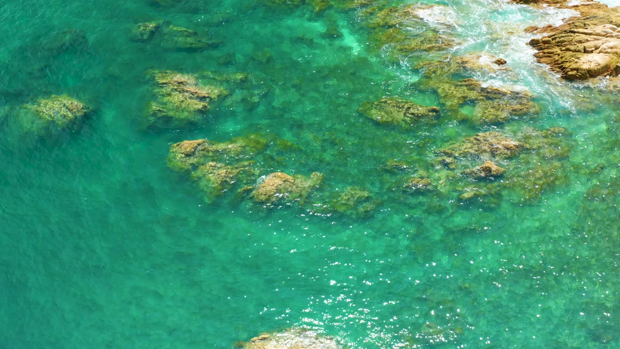 Clear teal water covers scattered reef rocks at Palmilla Beach, with sun flecks near the lower center and no visible people.