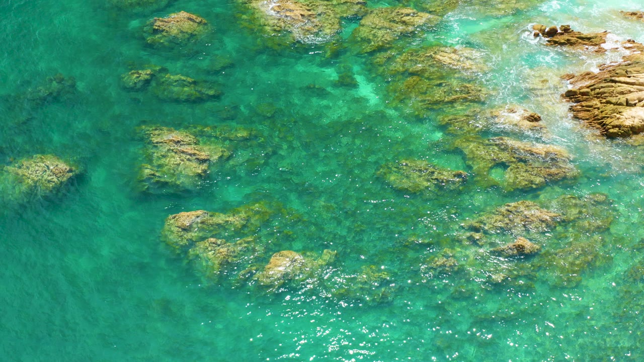Overhead frame at Palmilla Beach shows turquoise shallow water, scattered reef rocks, sun sparkle near the lower center, and no visible people.