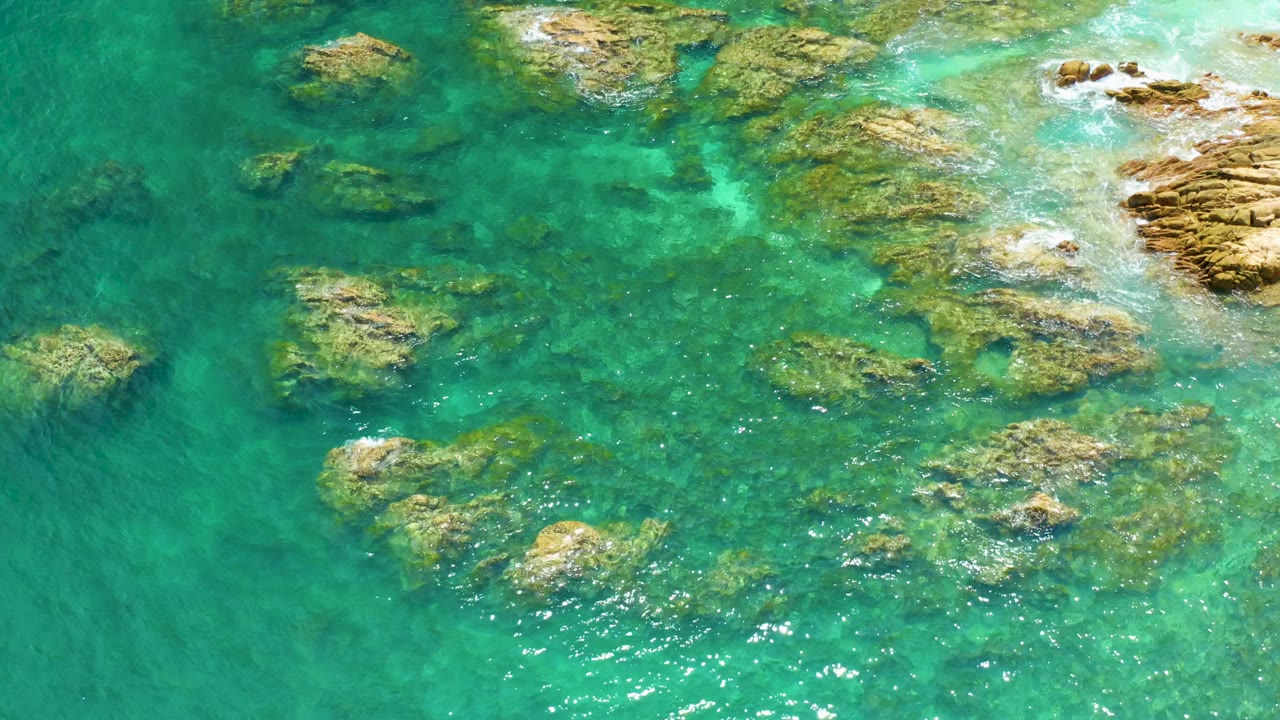 A top-down view at Palmilla Beach shows brighter turquoise water, exposed tan rocks near the right side, submerged reef shapes, and no visible people.