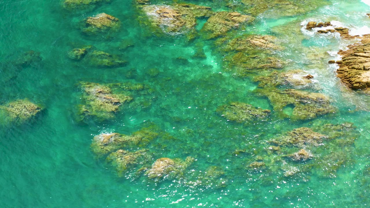 Palmilla Beach appears from above with green-blue water, reef clusters crossing the center, sun glints near the lower middle, and no visible people.