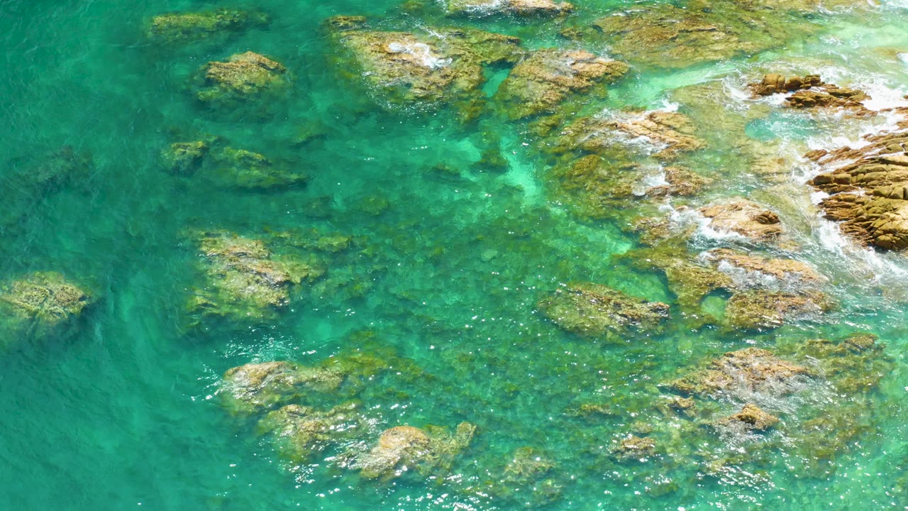One second into the Palmilla Beach clip, blue-green water covers uneven rocks while a small band of white wash moves near the upper right, with no visible people.