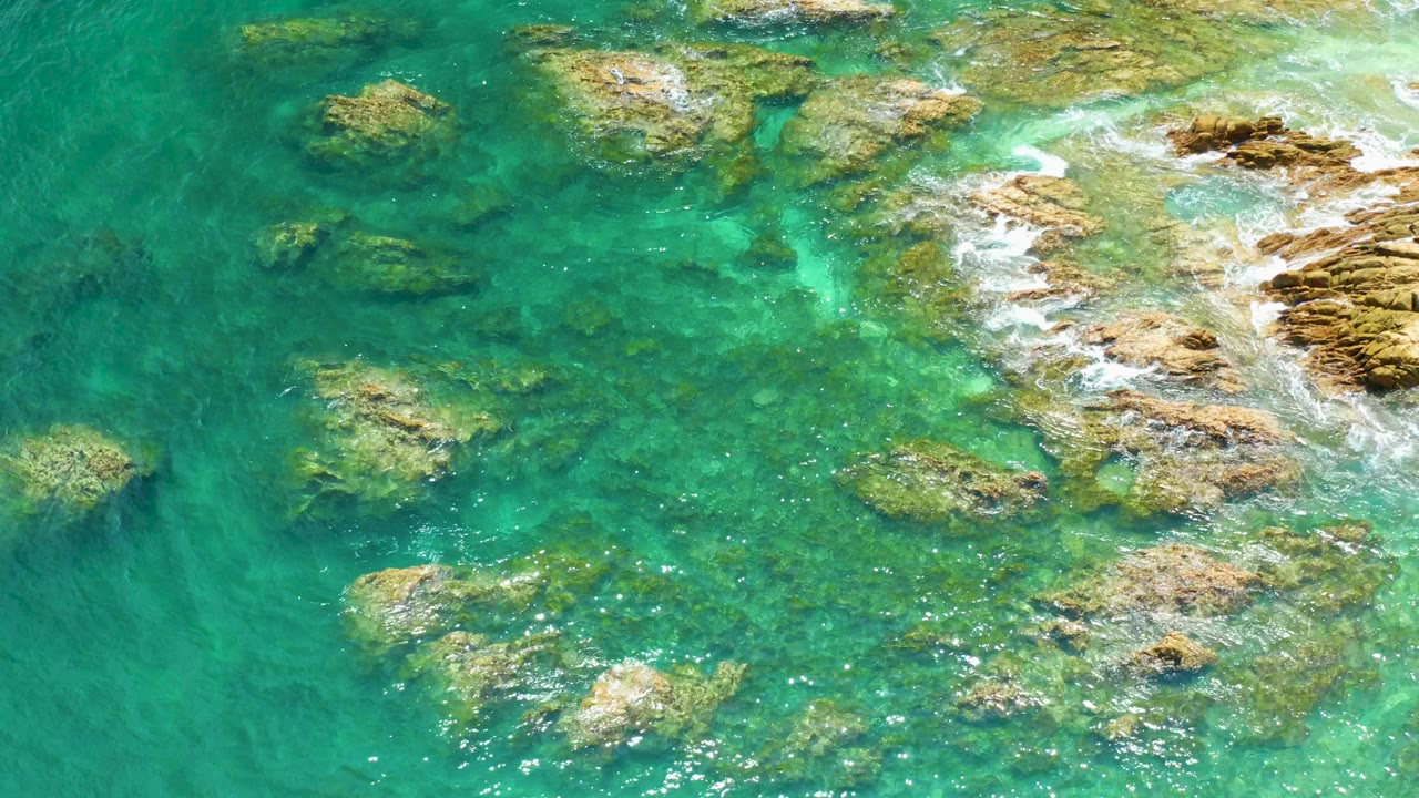Mid-clip overhead view at Palmilla Beach showing green-blue shallow water, several tan rock forms, a foamy wash line at right, and no visible people.