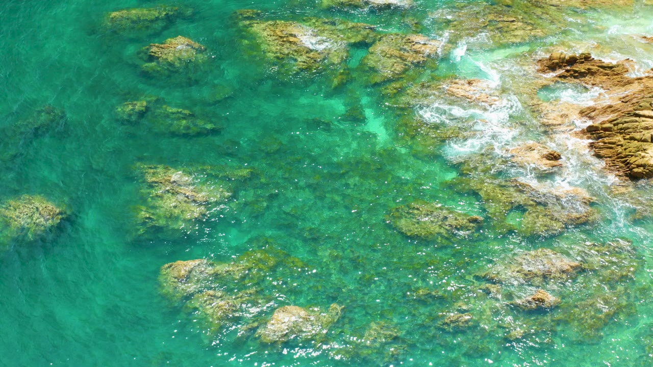 Aerial frame of Palmilla Beach showing turquoise water, scattered rocks, white surf at the right shoreline edge, and no visible people.