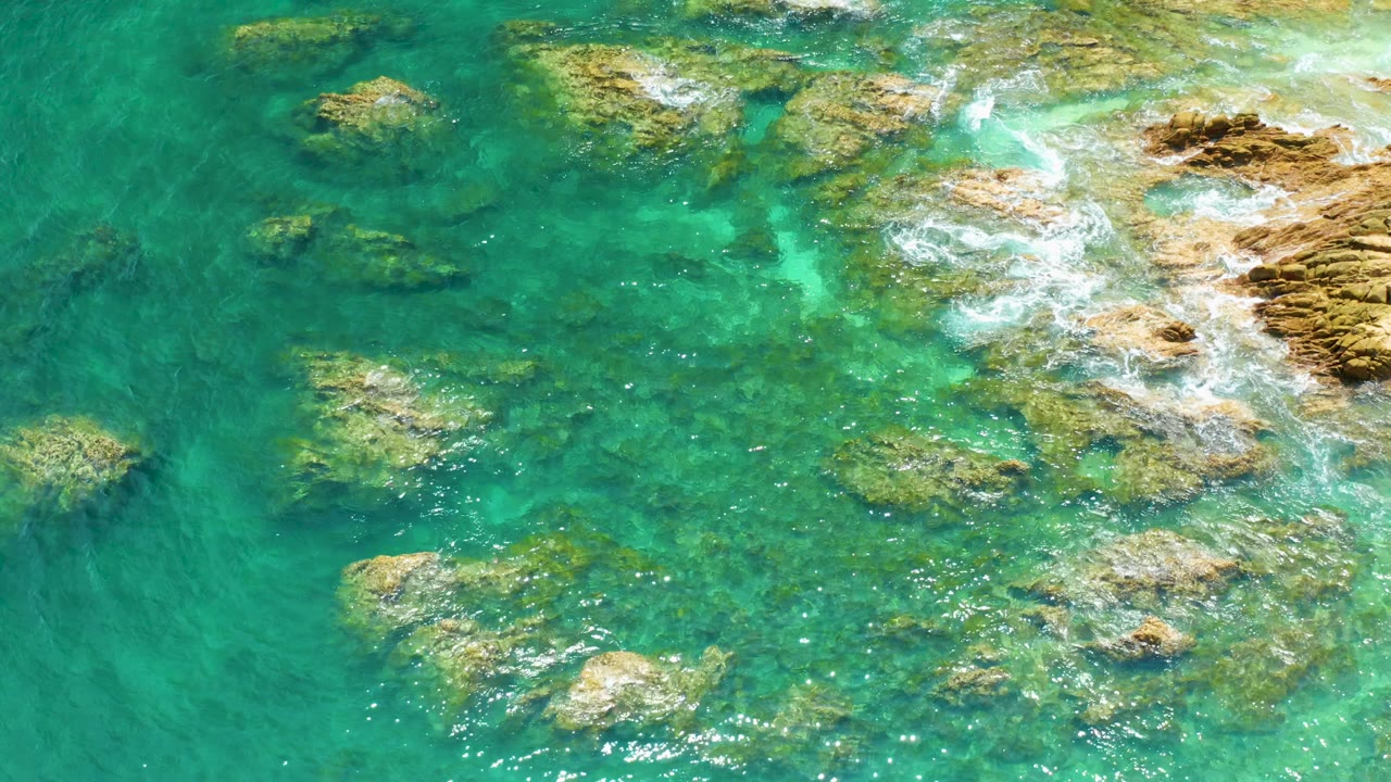 Aerial view at Palmilla Beach with green-blue shallows, several rock outcrops, foamy water along the right side, and no visible people.