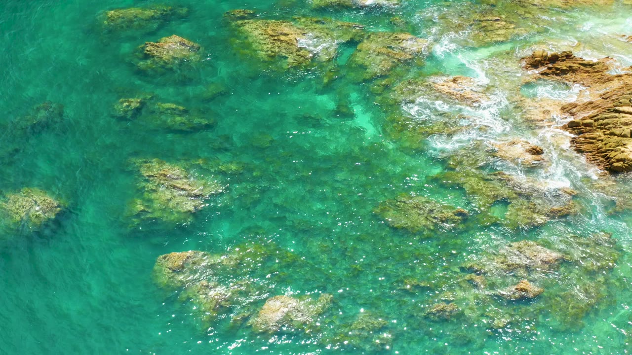 Palmilla Beach aerial frame with turquoise shallows, darker submerged rocks, bright whitewater near the right coastal rocks, and no visible people.