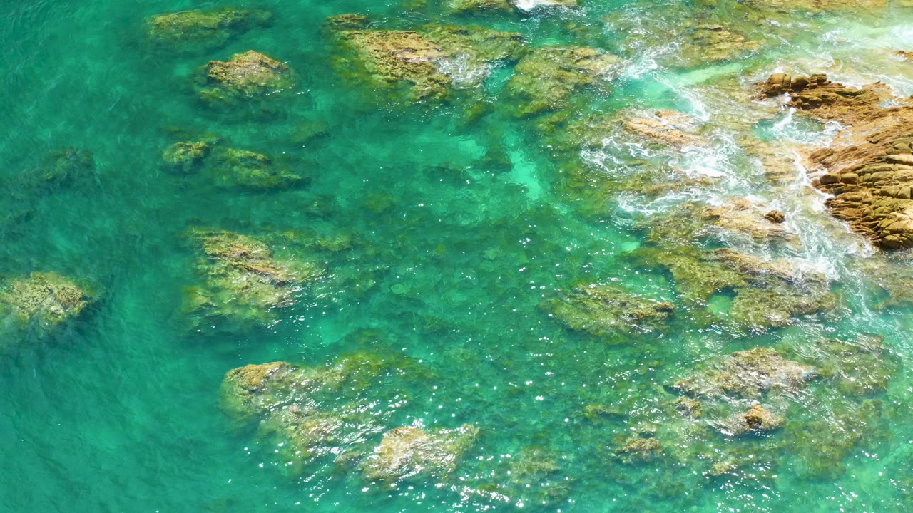 Aerial frame over Palmilla Beach where clear turquoise water surrounds scattered rocks and a band of white surf reaches in from the right, with no visible people.