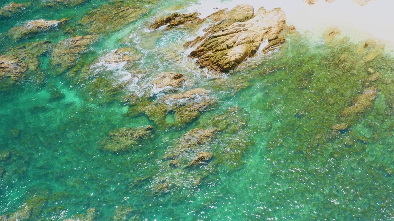Overhead view at Palmilla Beach, Mexico, showing turquoise shallow water, tan rocks near the top edge, small white foam lines, and no visible people.