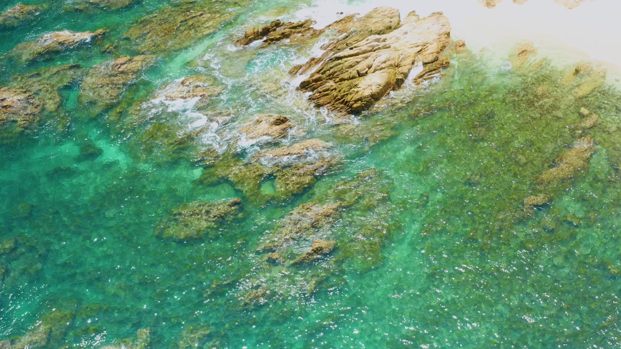 Palmilla Beach water seen from above, with the main tan rock cluster high in frame, smaller submerged rocks below it, bright reflections, and no visible people.