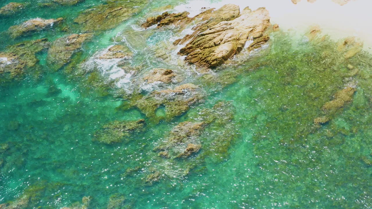 Aerial frame from Palmilla Beach showing clear ocean water over mottled rocks, white wash along the upper shoreline rocks, darker teal water at left, and no visible people.