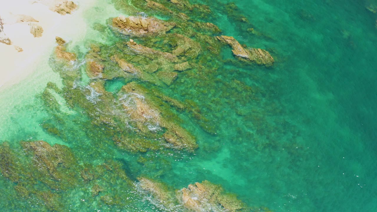 Overhead frame at Palmilla Beach shows pale sand at the upper left, tan submerged rocks across clear turquoise water, and no visible people.