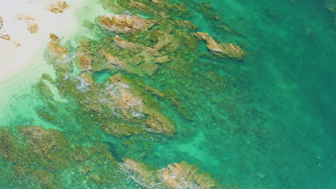 Aerial frame of clear green-blue shallows over clustered brown rocks at Palmilla Beach, with bright beach sand partly visible at upper left and no visible people.