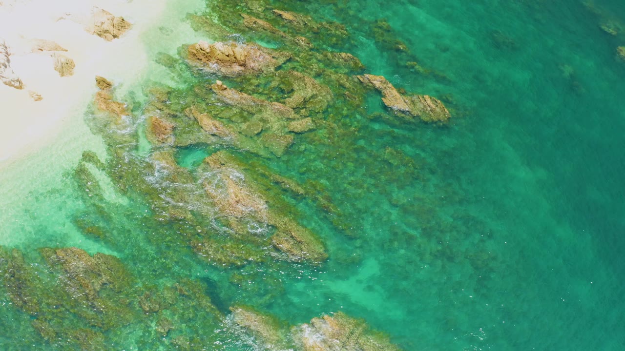 Top-down view at Palmilla Beach with pale sand in the upper left, a band of exposed and submerged rocks through the center, deeper turquoise water to the right, and no visible people.