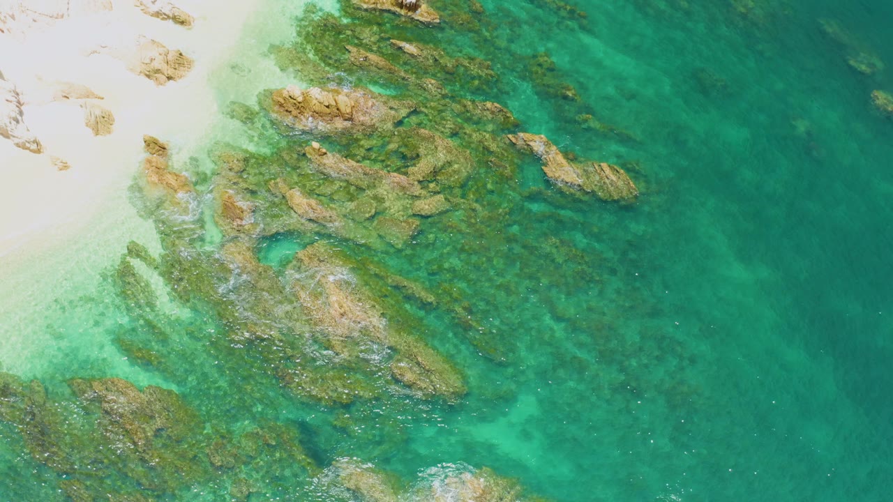 Overhead Palmilla Beach view with sunlit rock shelves under transparent water, pale sand at the left edge, faint wave foam around the rocks, and no visible people.