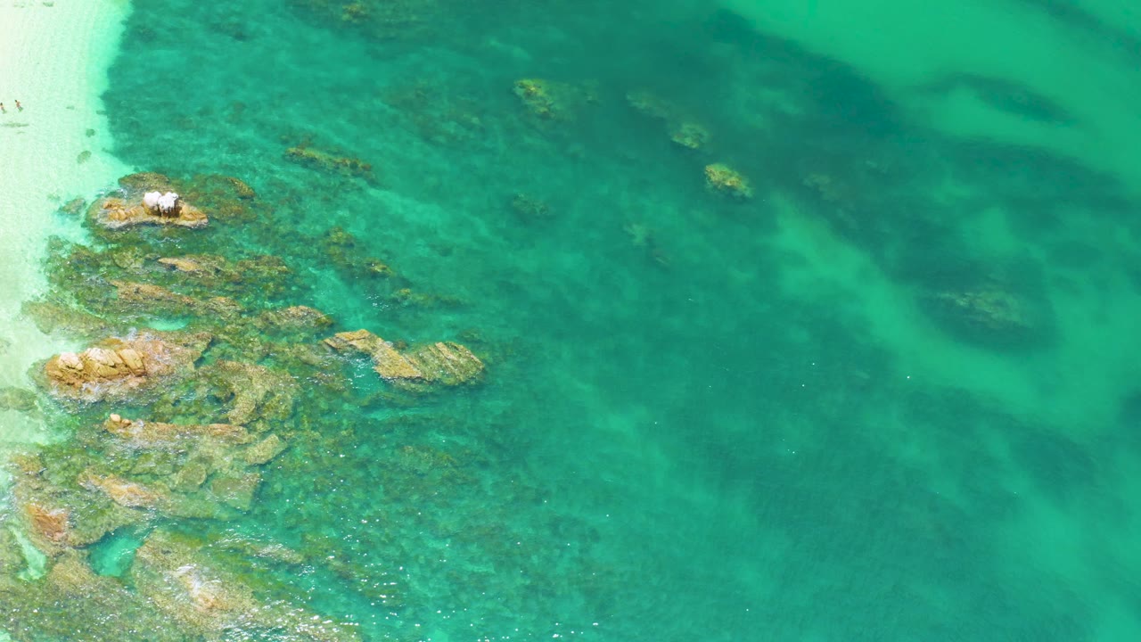 Overhead frame at Palmilla Beach showing clear turquoise water over submerged rocks, a pale sand edge at left, darker water patches to the right, and no visible people.