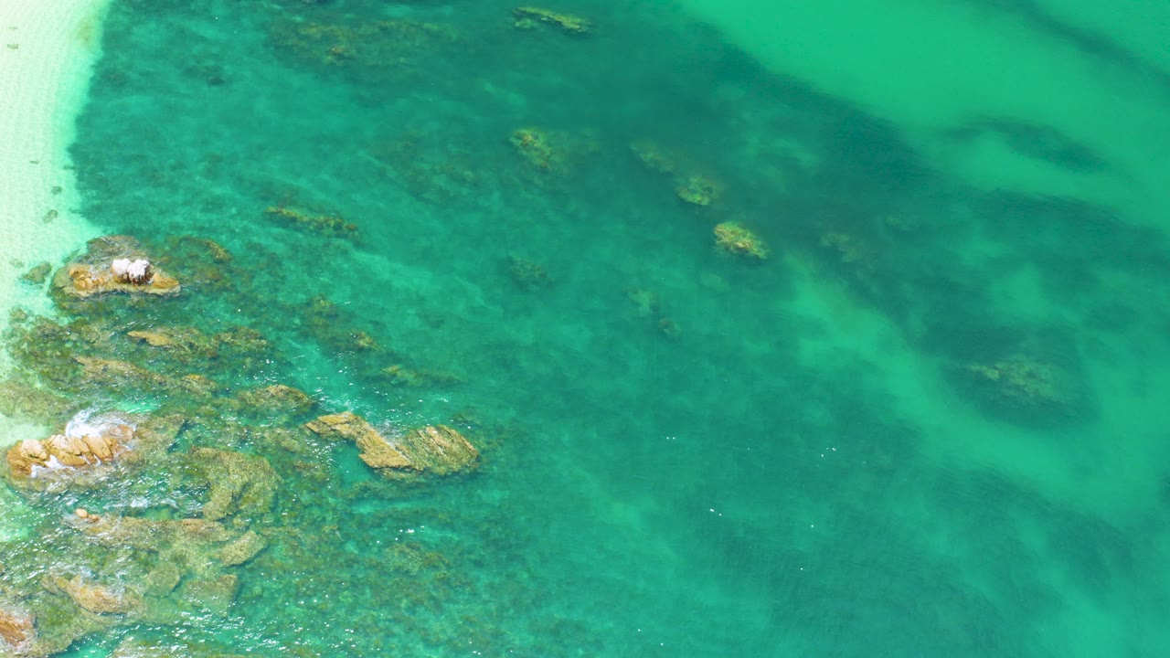 Overhead view at Palmilla Beach, Baja California Sur, showing clear turquoise water over scattered rocks, a pale sand edge at left, and no visible people.