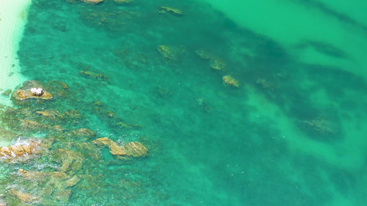 Overhead Palmilla Beach view with tan rocks under clear turquoise water, faint white surf around the left-side rocks, and no visible people.