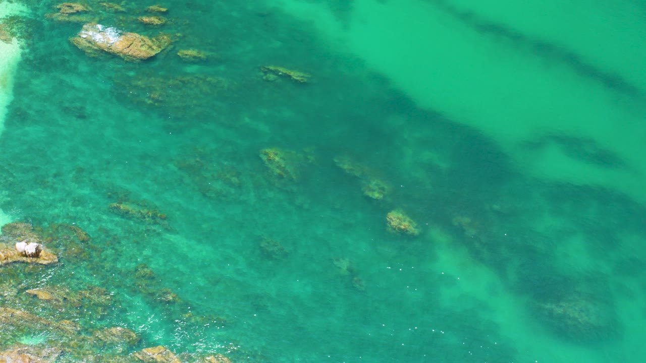 Overhead view of turquoise water at Palmilla Beach, Baja California Sur, with scattered submerged rocks, a small wave-washed outcrop near the lower left, and no visible people.