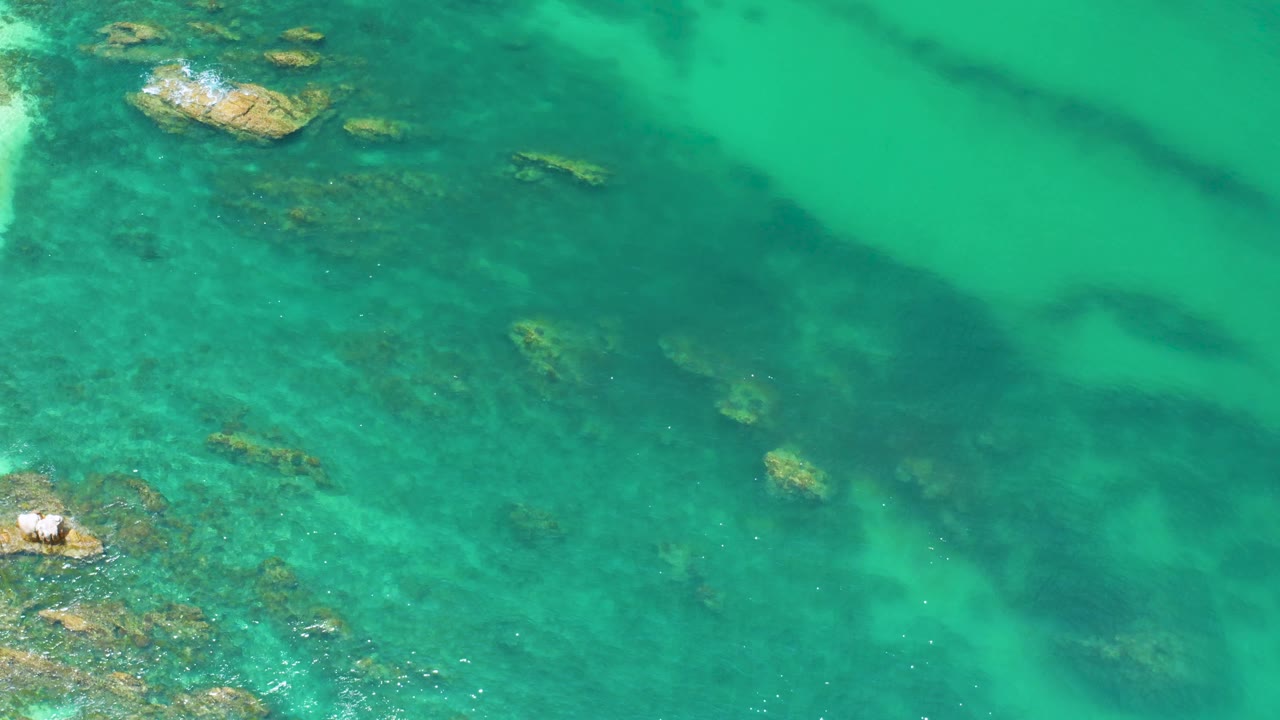 Clear green-blue Palmilla Beach water shows submerged rock detail, a small foamy rocky outcrop along the left edge, and no visible people.