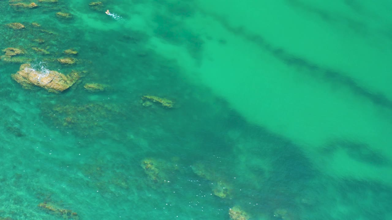 Top-down Palmilla Beach water view with the main rock cluster higher in frame, darker submerged bands crossing the water, and no visible people.