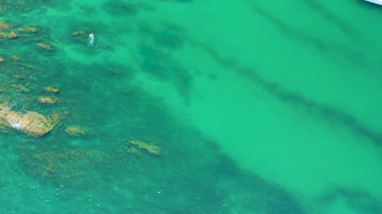 Aerial view over Palmilla Beach water with clustered reef patches on the left and smooth green water spreading across the right side, no visible people.
